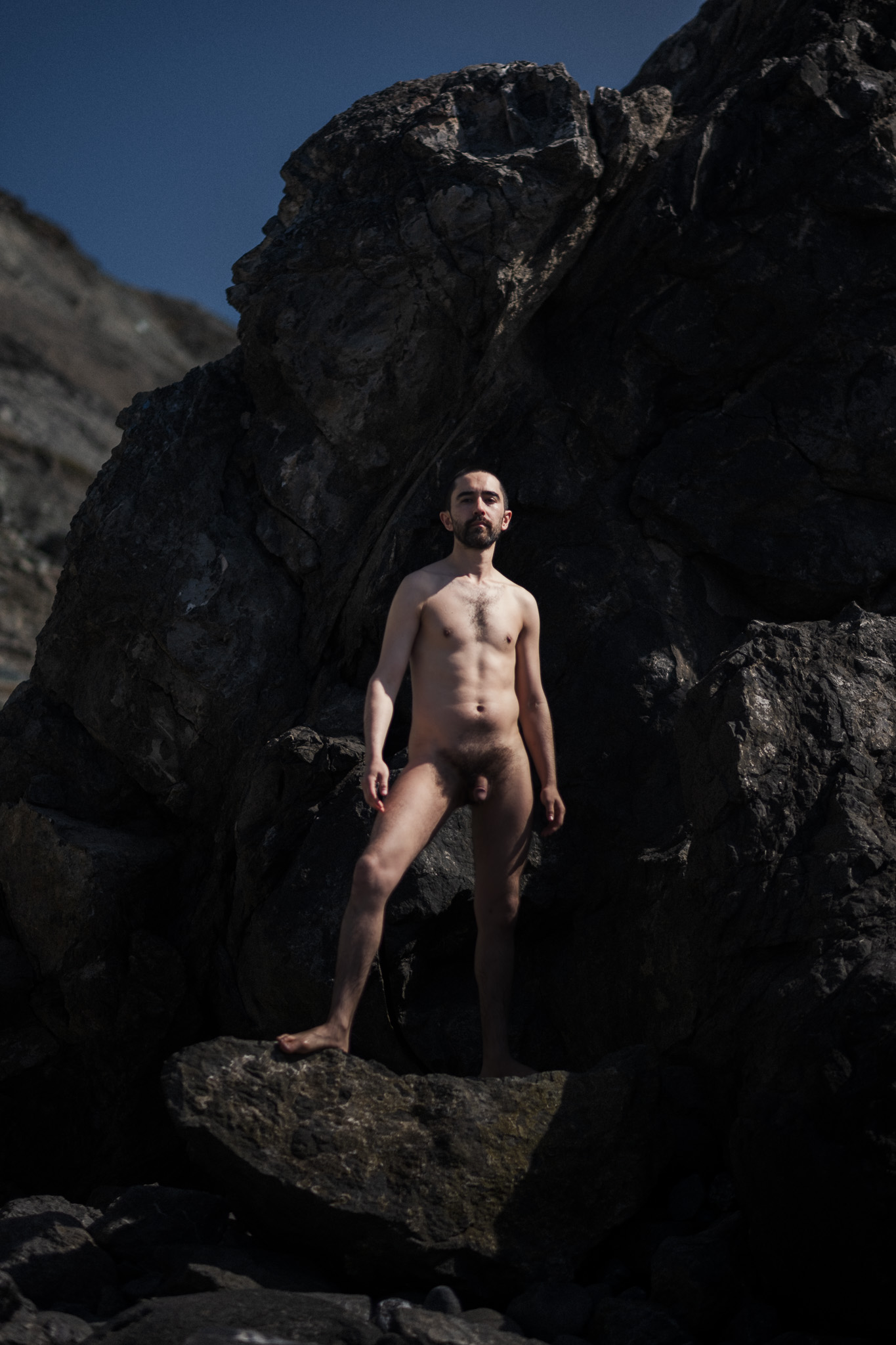 Nude self-portrait standing against dark coastal rocks